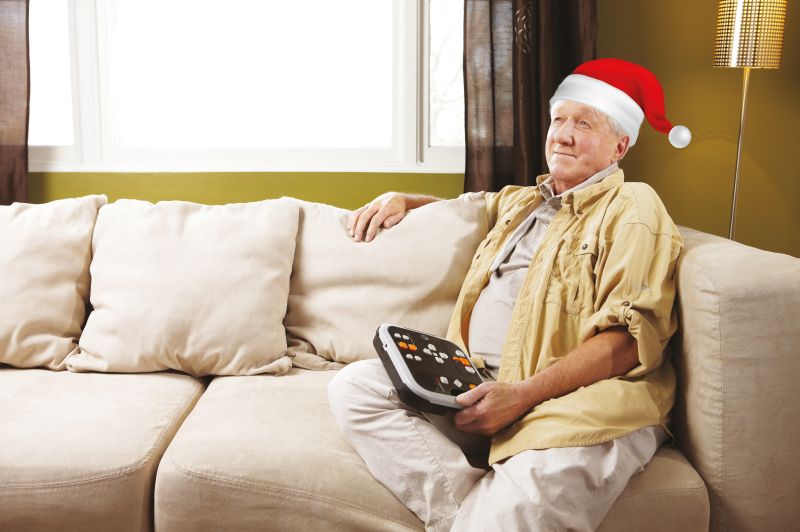 Mann sitzt auf dem Sofa und hört ein Hörbuch mit seinem Victor Reader - dabei trägt er eine weihnachtliche Mütze